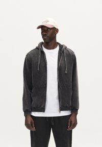 Gray hoodie with front pockets and zipper, featuring a relaxed fit and an adjustable hood. Worn over a white T-shirt, matching dark pants.