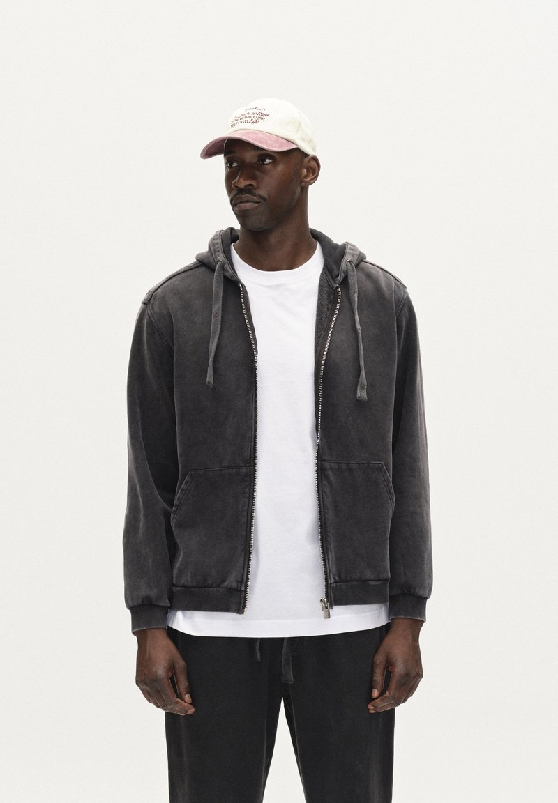 Gray hoodie with front pockets and zipper, featuring a relaxed fit and an adjustable hood. Worn over a white T-shirt, matching dark pants.