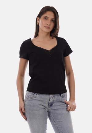 Femme portant un haut noir à manches courtes avec col en V et détails en dentelle, ainsi qu'un jean gris clair, debout avec une main dans la poche sur un fond uni.