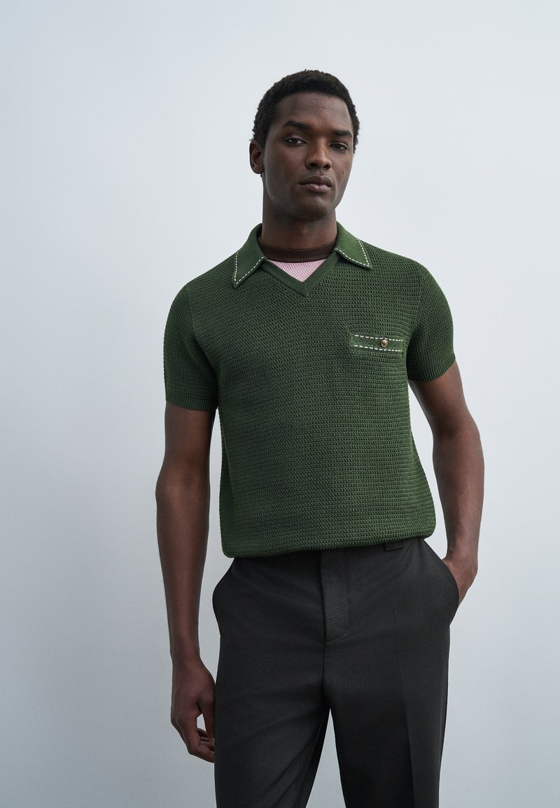 Άνδρας που φοράει σκούρο πράσινο υφασμάτινο πόλο με κοντά μανίκια και τσέπη στο στήθος, καθώς και μαύρο παντελόνι, στέκεται μπροστά σε απλό ανοιχτό γκρι φόντο.