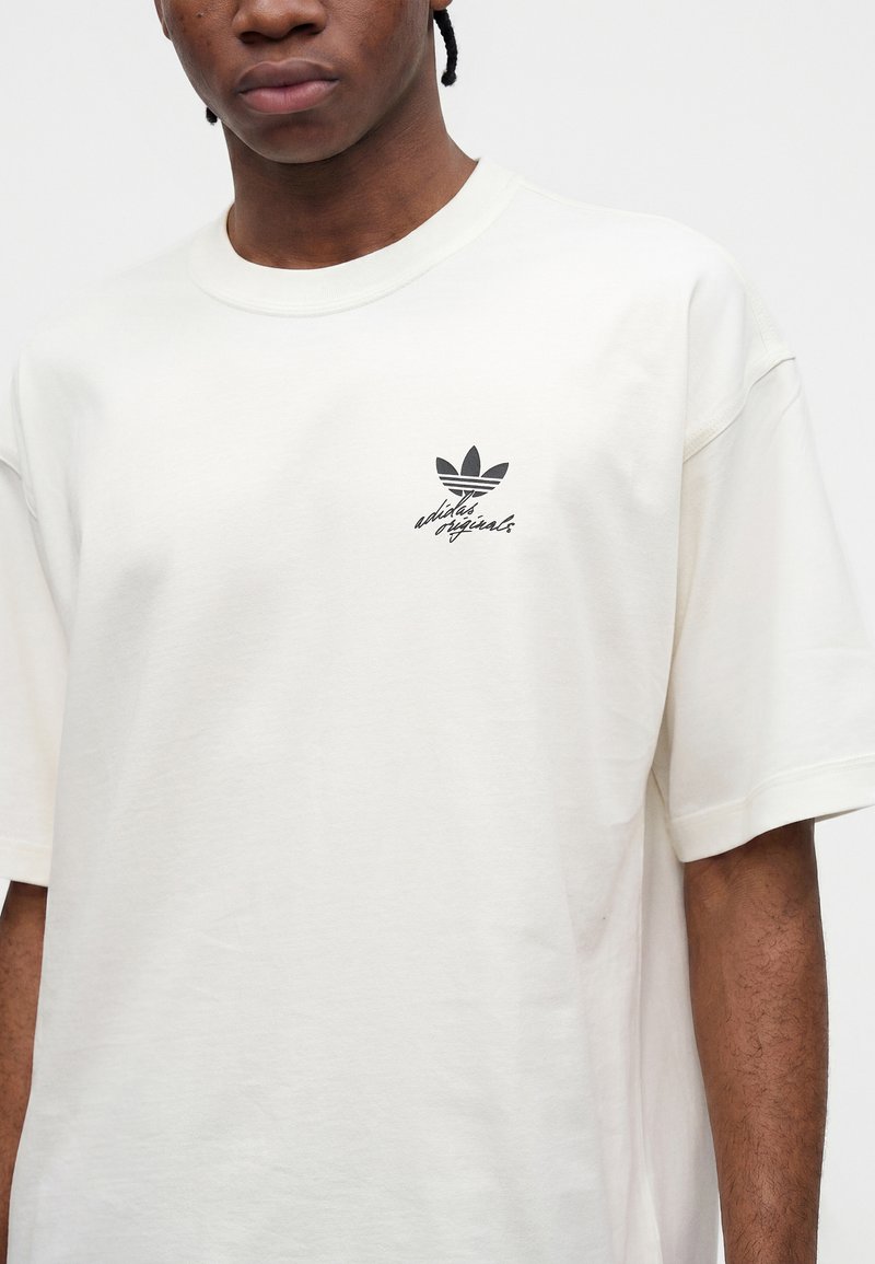 Jeune personne portant un t-shirt blanc Adidas Originals avec un logo trèfle noir et un texte sur la poitrine gauche sur un fond uni.