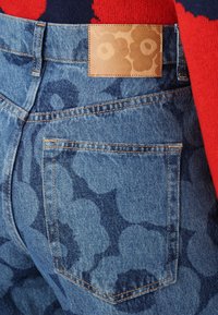 Jeans aus Denim mit einem blauen Blumenmuster, ausgestattet mit einem braunen rechteckigen Lederpatch und orangefarbenen Nähten, die das Design der Gesäßtasche hervorheben.