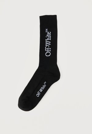 Calcetín negro acanalado hasta el tobillo con el texto "Off-White™" en blanco, colocado verticalmente en la pantorrilla y horizontalmente en la planta del pie.