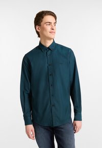 Donkerblauwe button-up shirt met lange mouwen en een puntige kraag, zwarte knopen, en een klein logo op de borst. Glad, lichtgewicht stof.