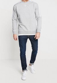 Šedá mikina s třemi bílými pruhy na ramenou, v kombinaci s tmavými džíny a bílými teniskami. Jednoduchý design, žebrované manžety a lem.