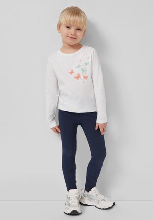 Young child with blonde hair wearing a white long-sleeve shirt with pastel butterflies, navy leggings, and white sneakers standing against a wall.