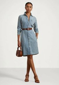 Kvinna i en knappad denimklänning med ett brunt bälte, som håller en brun handväska och har på sig bruna klacksandaler, stående mot en neutral bakgrund.