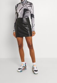 Schwarzer Leder-Minirock mit glatter Textur, kombiniert mit einem grauen, abstrakt gemusterten Langarmoberteil und bunten Sport sneakers.