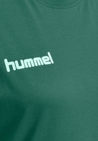 Camiseta verde de algodón con cuello redondo y textura suave, con un logo blanco impreso "hummel" en el frente.