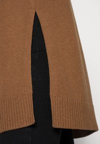 Brązowy sweter z dzianiny z bocznymi rozcięciami, o prążkowanym wykończeniu i miękkiej teksturze. W zestawieniu z czarnym materiałem pod spodem, podkreślającym kontrast kolorystyczny.