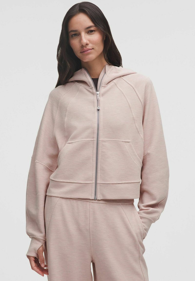 Hanorac scurt roz, cu un aspect texturat, cu fermoar în față și buzunare mari. Are o glugă și umeri ușor lăsați.