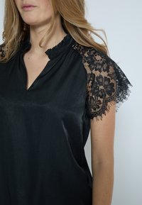 Sort kortærmet top med en glat stofkrop, lace detaljer på skuldrene og en rynket krave.