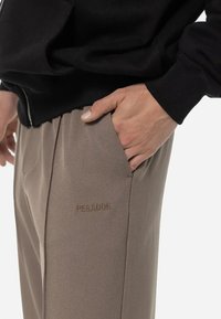 Pantalons beiges avec poches latérales et finition texturée, arborant un logo en relief "PEGADOR" sur la cuisse gauche. Porté avec un pull noir.