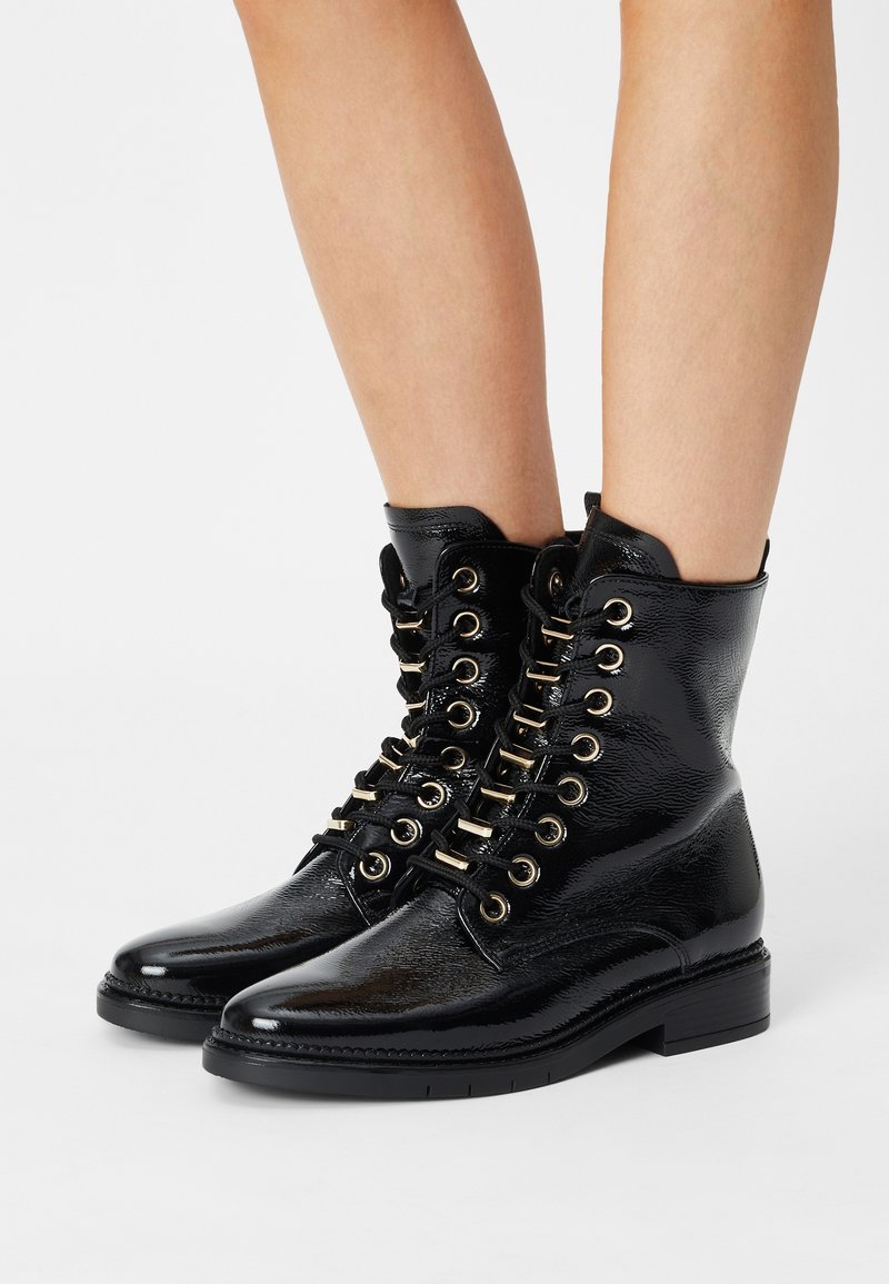 Bottes noires brillantes à chevilles avec œillets dorés et lacets noirs, portées sur des jambes nues, avec un talon bas et une finition texturée.