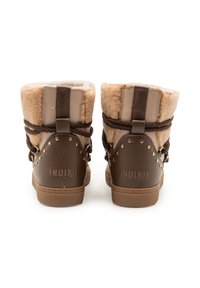 Een paar bruine en beige met bont gevoerde winterlaarzen met gevlochten banden en gouden studs, van achteren gezien op een witte achtergrond.