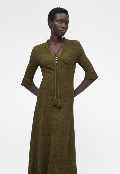 Γυναίκα που φοράει ελιά-πράσινο, φόρεμα μήκους έως το γόνατο με δαντελένιο σχέδιο, με μανίκια τριών τετάρτων και κορδόνια με φούντες στο ντεκολτέ, πάνω σε λευκό φόντο.