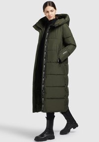 Donna che indossa un lungo cappotto imbottito verde oliva con cappuccio, leggings neri e stivali neri fino a metà polpaccio, in piedi contro uno sfondo semplice.