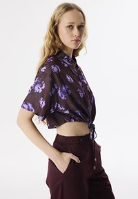 Korte mouwen, cropped shirt in donkerbruin met een paars bloemenpatroon; heeft een kraag en een elastische onderrand, gecombineerd met hoog-waist bordeauxrode broek.