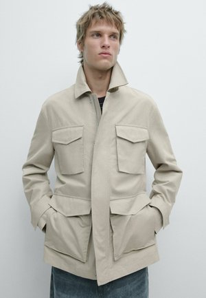 Junger Mann mit zerzaustem blondem Haar, trägt eine beigen Jacke mit vier großen Fronttaschen und hat die Hände in den Seitentaschen, blickt nach rechts.