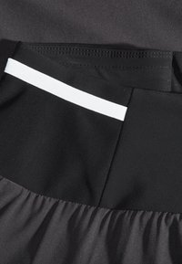Svart textil med en slät, glänsande yta och nätpaneler, med en vit rand som accentuering på midjebandet. Rynkad nederkant.