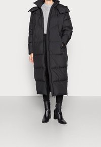 Svart puffjacka med hög krage, dragkedja framtill och sidofickor, med ett kviltat mönster. Bärs över en vit mönstrad topp och svarta byxor.