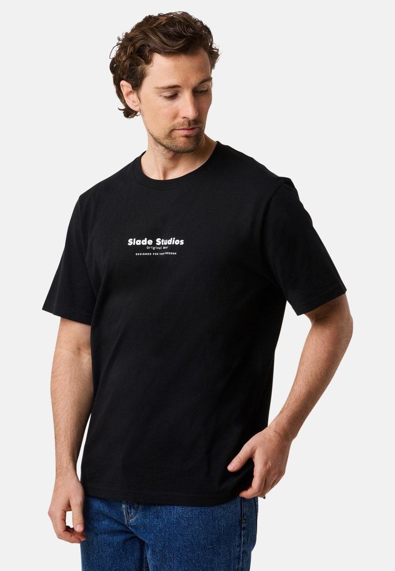 Zwarte katoenen t-shirt met korte mouwen, met witte tekst "Slade Studios" en "ONTWORPEN VOOR JOUW VRIJHEID" op de voorkant.