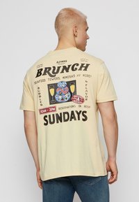 Blond mees kerge kollase T-särgiga, mille seljal on brunchi graafika, kus on kujutatud mereandide torne, mimosaid ja broneeringute teksti.