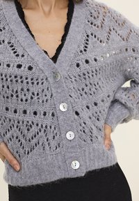 Grau gestrickte Strickjacke mit V-Ausschnitt, die ein Spitzenmuster, einen gerippten Saum und fünf runde, schimmernde Knöpfe aufweist. Weicher Stoff und lockere Passform.