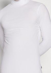 Mann in einem enganliegenden weißen Langarm-Turtleneck-Shirt, der vor einem neutralen Hintergrund steht.