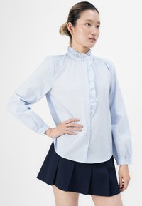 Kvinne iført en lyseblå stripete bluse med volanger og et marineblått plisseet skjørt, poserer med en hånd på hoften mot en hvit bakgrunn.