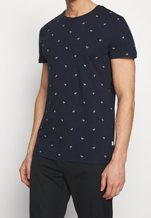 Mies pukeutuneena tummansiniseen lyhythihaiseen t-paitaan, jossa on pieniä valkoisia paperilentokonekuvioita, yhdistettynä mustiin housuihin, seisoo näkyvällä yhdellä kädellä.