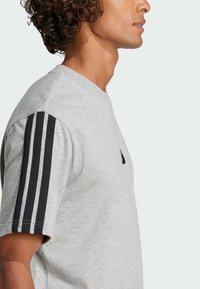 Šedé tričko s krátkym rukávom vyrobené z jemnej látky, so čiernymi trojpruhovými akcentmi na pleciach a malým logom na hrudi.