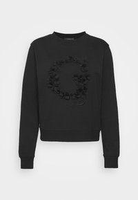 Sudadera negra con cuello redondo, con un diseño floral texturizado que forma una "G" y el nombre de la marca "Guess" en cursiva en la parte inferior.