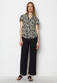 Schwarze kurzärmlige Bluse mit einem floralen cremefarbenen Muster, mit einer durchgehenden Knopfleiste und einem lockeren Schnitt. Kombiniert mit maßgeschneiderten schwarzen Hosen und Sandalen.
