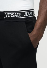 Κοντινό πλάνο ενός ατόμου που φοράει μαύρο παντελόνι με λάστιχο μέσης με την ένδειξη "Versace Jeans Couture" και το χέρι ξεκουρασμένο στην τσέπη.