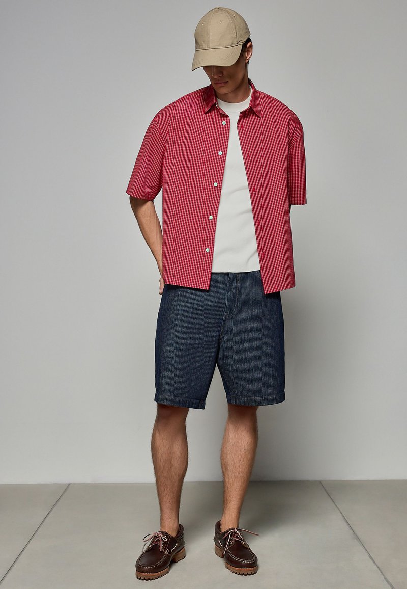 Cămașă scurtă cu maneci scurte în carouri roșii și albe, maieu alb, pantaloni scurți din denim și pantofi maro. Accesoriile includ o șapcă de baseball bej.