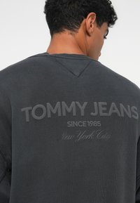 Felpa grigia scura con grande testo in rilievo "TOMMY JEANS" sulla schiena, con "SINCE 1985" e "New York City" stampati in dimensioni più piccole sottostante.