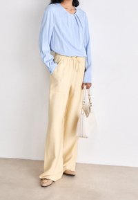 Světle modrá halenka s dlouhým rukávem a záhybem, kombinovaná s krémovými širokými kalhotami a béžovou kabelkou. Světlá látka, volný střih.