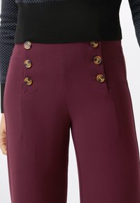 Pantaloni bordeaux in tessuto testurizzato, con quattro bottoni dorati sui lati e tasche frontali, abbinati a un maglione nero aderente.