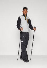 Fleece pullover in lichtgrijs en zwart met een ritskraag, gele ritssluitingsdetails, zwarte broek en trekkingstokken, met reflecterende elementen.