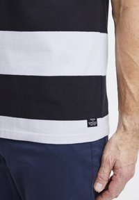 Melna un baltā svītrainā t-kreklā, kas izgatavots no kokvilnas, ar apaļu izgriezumu un nelielu etiķeti apakšmalā. Apvienots ar tumši zilām biksēm.