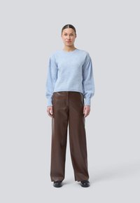 Hellblauer gestrickter Pullover mit puffigen Ärmeln, kombiniert mit weiten braunen Lederhosen und schwarzen Schuhen. Schlichter Hintergrund.