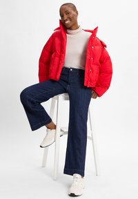 Rød puffjakke med høy krage, kombinert med en tettsittende kremfarget genser med høy hals og brede mørk denimjeans. Hvite sneakers med svarte detaljer.