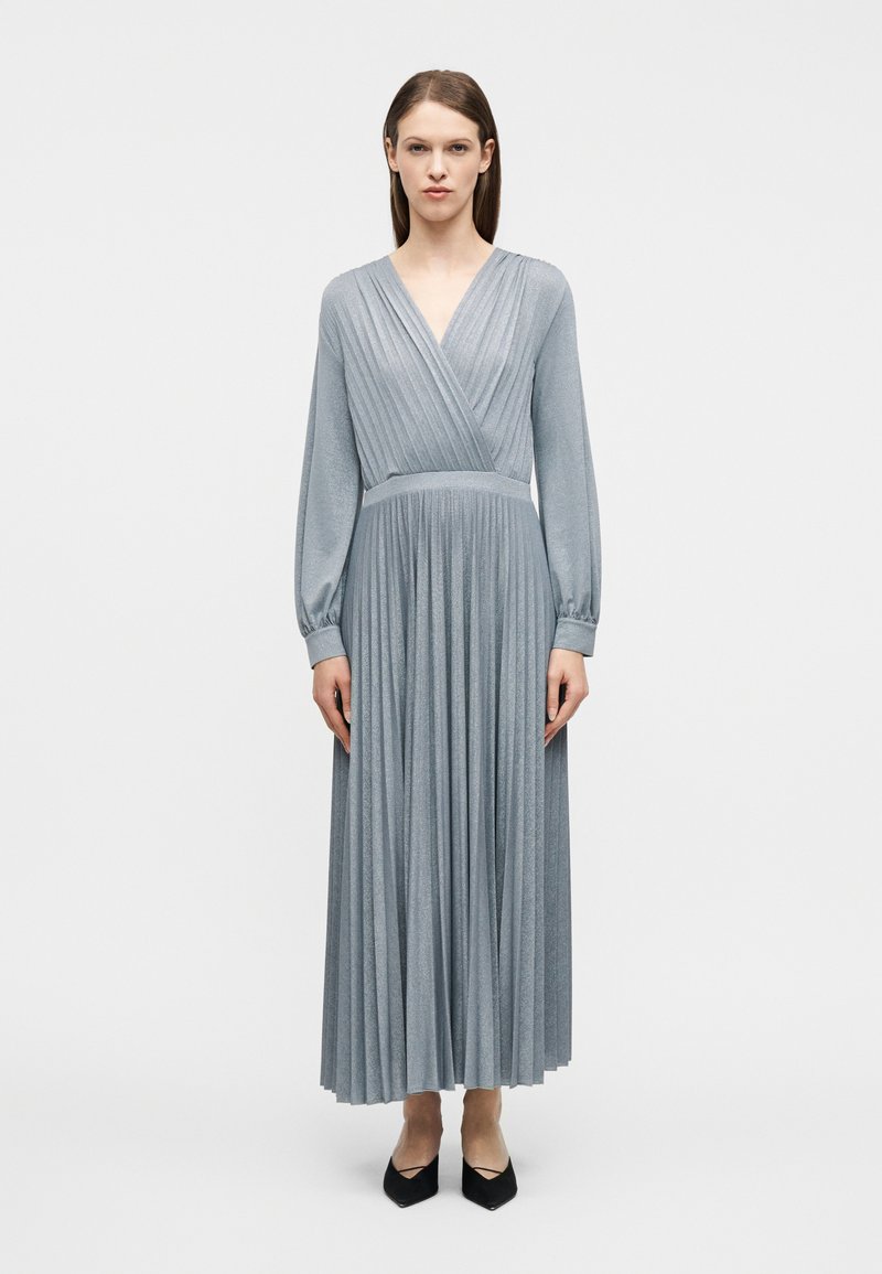 Γυναίκα που στέκεται φορώντας μακρυμάνικο, πλισέ, ασημί-γκρι μάξι φόρεμα με μαύρα παπούτσια μυτερά σε ένα απλό λευκό φόντο.