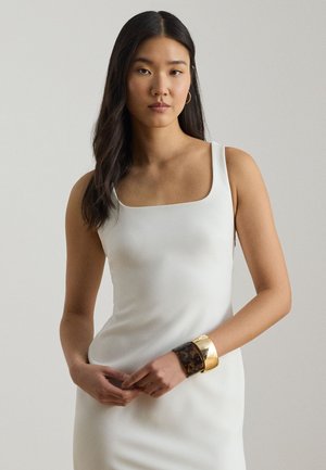 Mladá žena s dlhými tmavými vlasmi, oblečená v bielych bezrukávových šatách, so zlatými kruhovými náušnicami a dvoma širokými náramkami na zápästí.