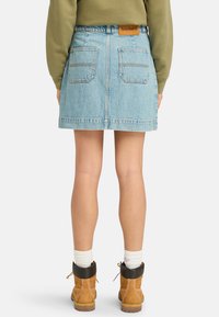 Falda de mezclilla azul claro con dos bolsillos traseros y una etiqueta de cuero marrón. El modelo la combina con botas de tobillo marrones y calcetines blancos.