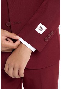 Manga de chaqueta de traje marrón con una etiqueta de tela blanca que dice "OPPO SUITS." Botones visibles y puño con forro blanco. Textura suave.