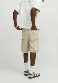 Muž oblečený v béžových cargo šortkách a bílých teniskách, stojící s jednou rukou v zadní kapse na světlém jednobarevném pozadí.