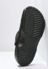 Semelle de sandale Crocs noire montrant le motif de la semelle et les marquages de taille, positionnée verticalement contre un fond blanc.