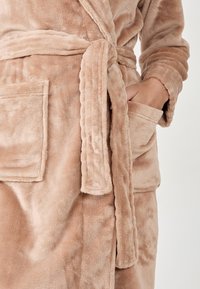 Accappatoio in morbida peluche color marrone chiaro con cintura annodata, texture soffice, due tasche frontali e colletto scialle. Il tessuto appare spesso e accogliente.